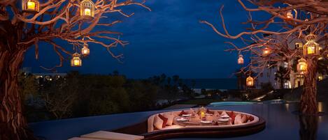 An outdoor dining area with a curved bench, lanterns, and a view of the ocean at night.