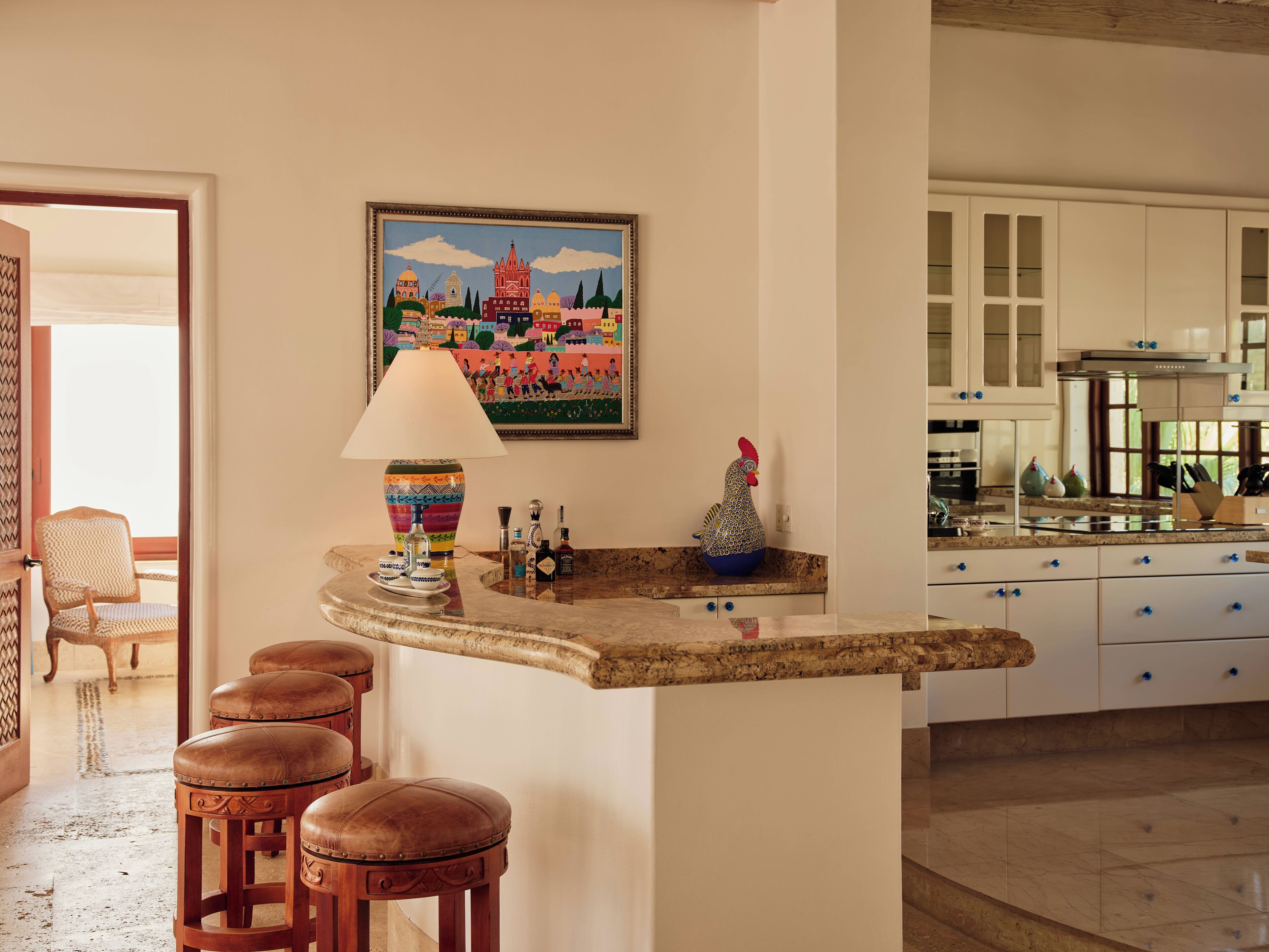 A kitchen with white cabinets, a marble countertop, and a colorful painting of a cityscape. There are stools and a lamp on the counter.