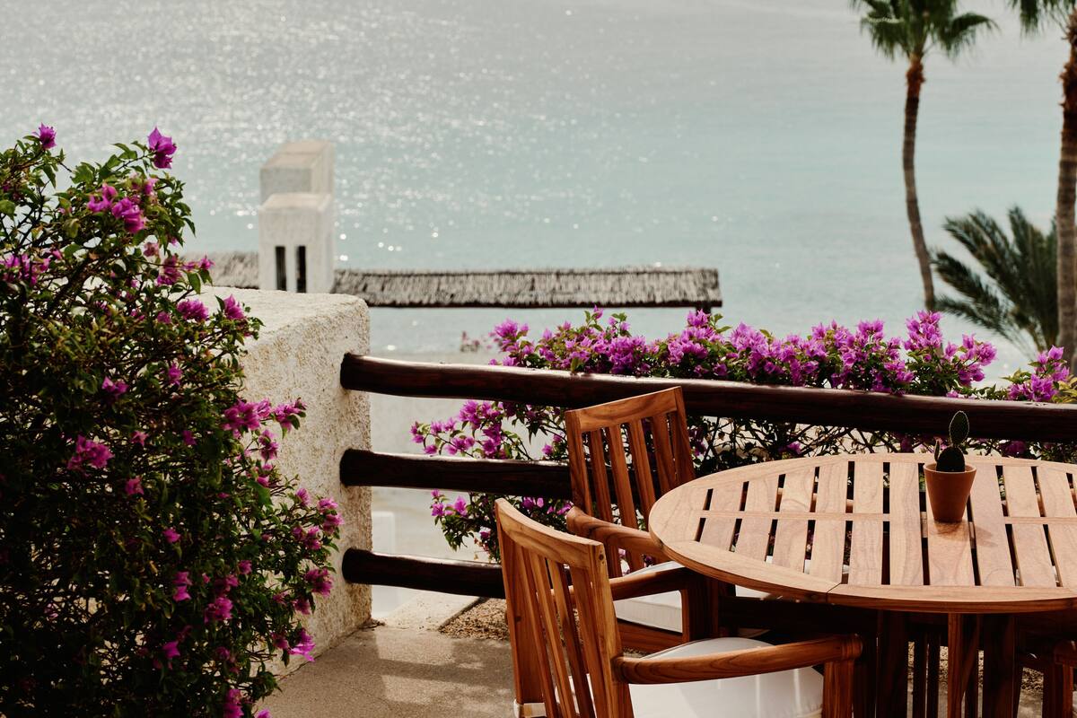 A wooden table and chairs on a balcony overlooking a body of water, with purple flowers and a palm tree in the background.