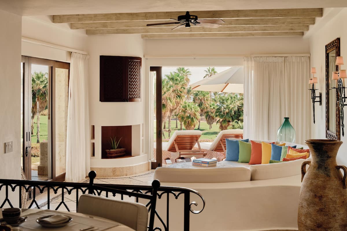 A room with a dining table, chairs, a sofa with colorful cushions, a large vase, and a view of palm trees outside.