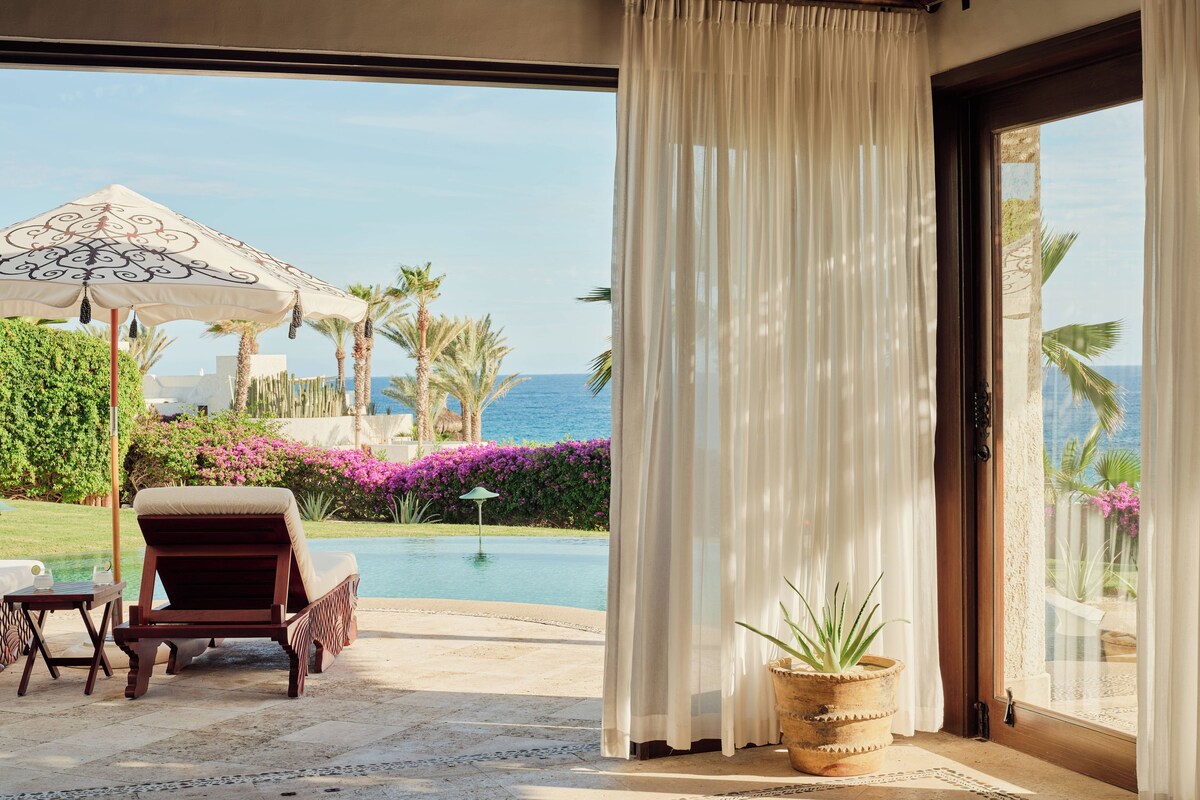 A room with a view of a pool and the sea, featuring a sliding glass door, a potted plant, and outdoor furniture.