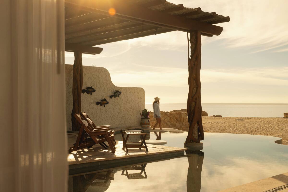 A beachside infinity pool with wooden chairs, a covered seating area, and a person walking.