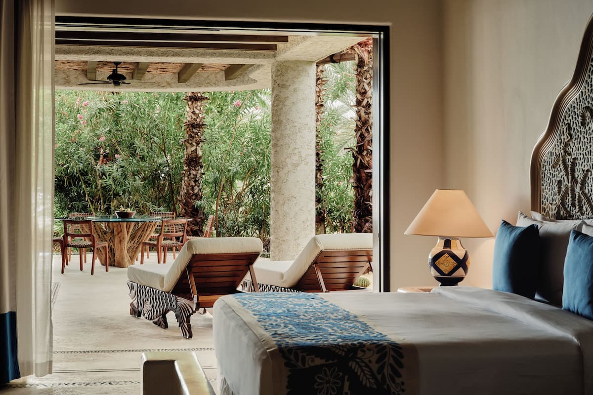 A bedroom with a bed, a nightstand, a lamp, and a view of an outdoor garden area with a table and chairs.