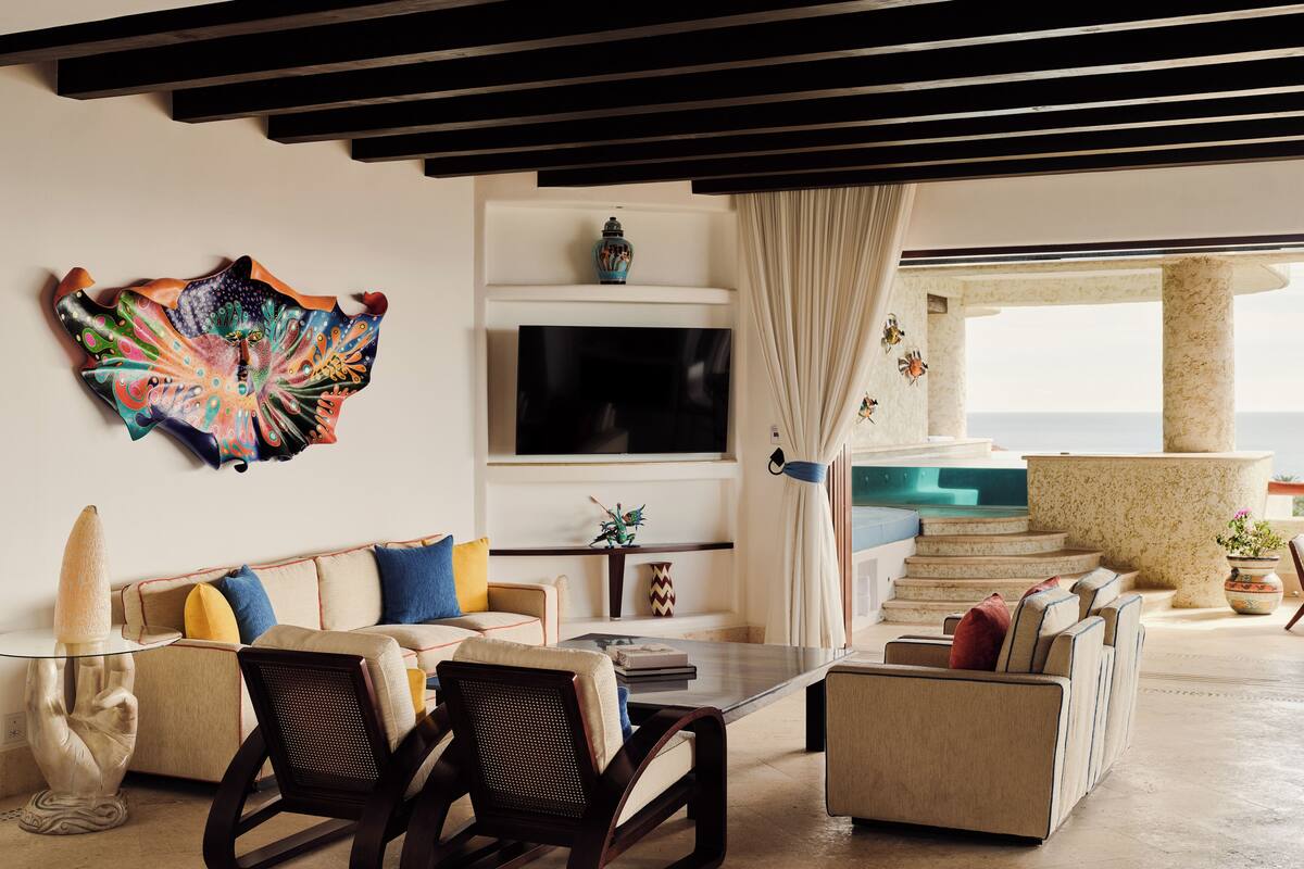 A living room with a sofa, chairs, a TV, a decorative wall piece, and a view of the ocean.