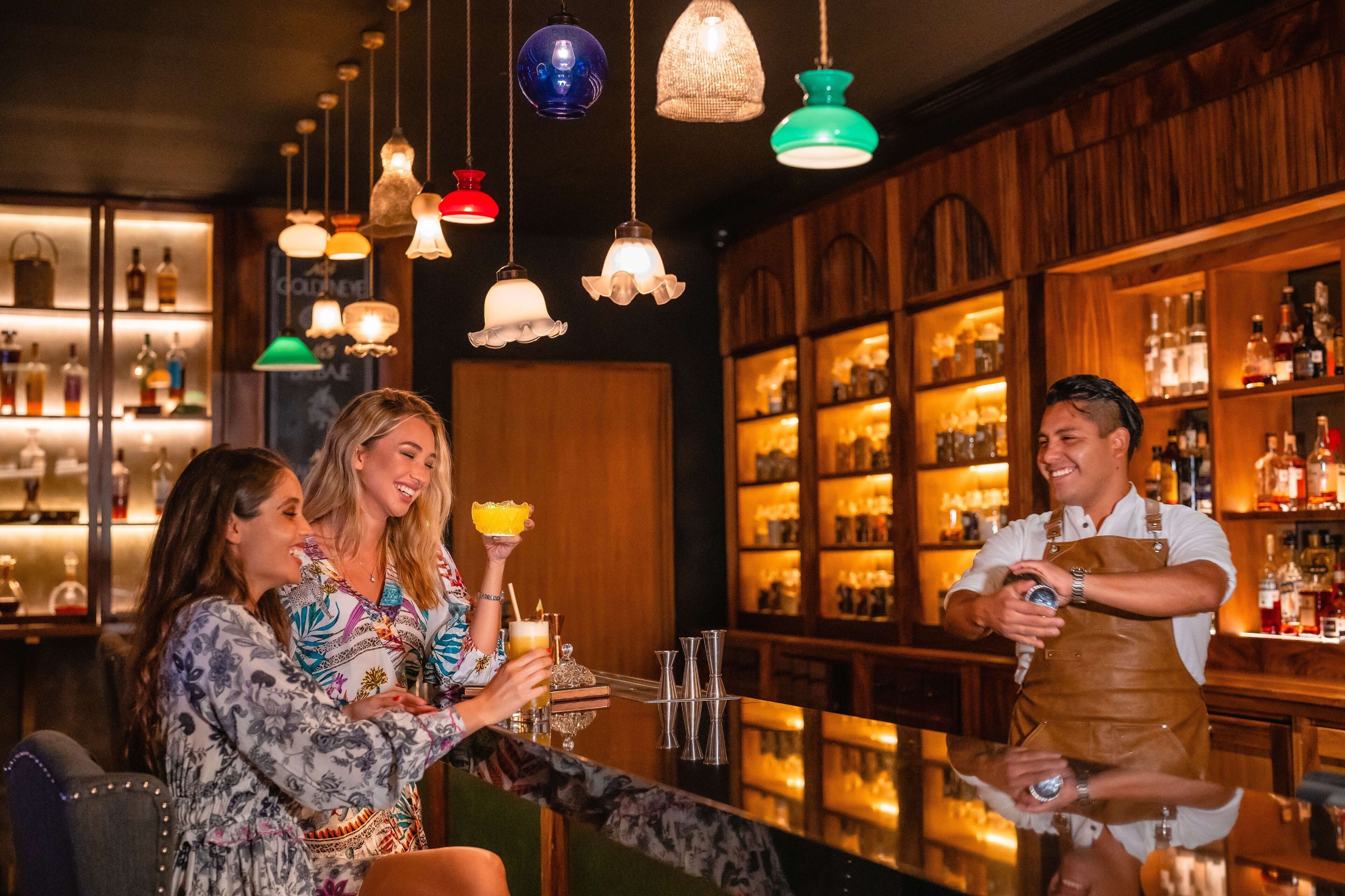 A bar area with wooden shelves stocked with bottles, hanging pendant lights, and a bartender preparing drinks.