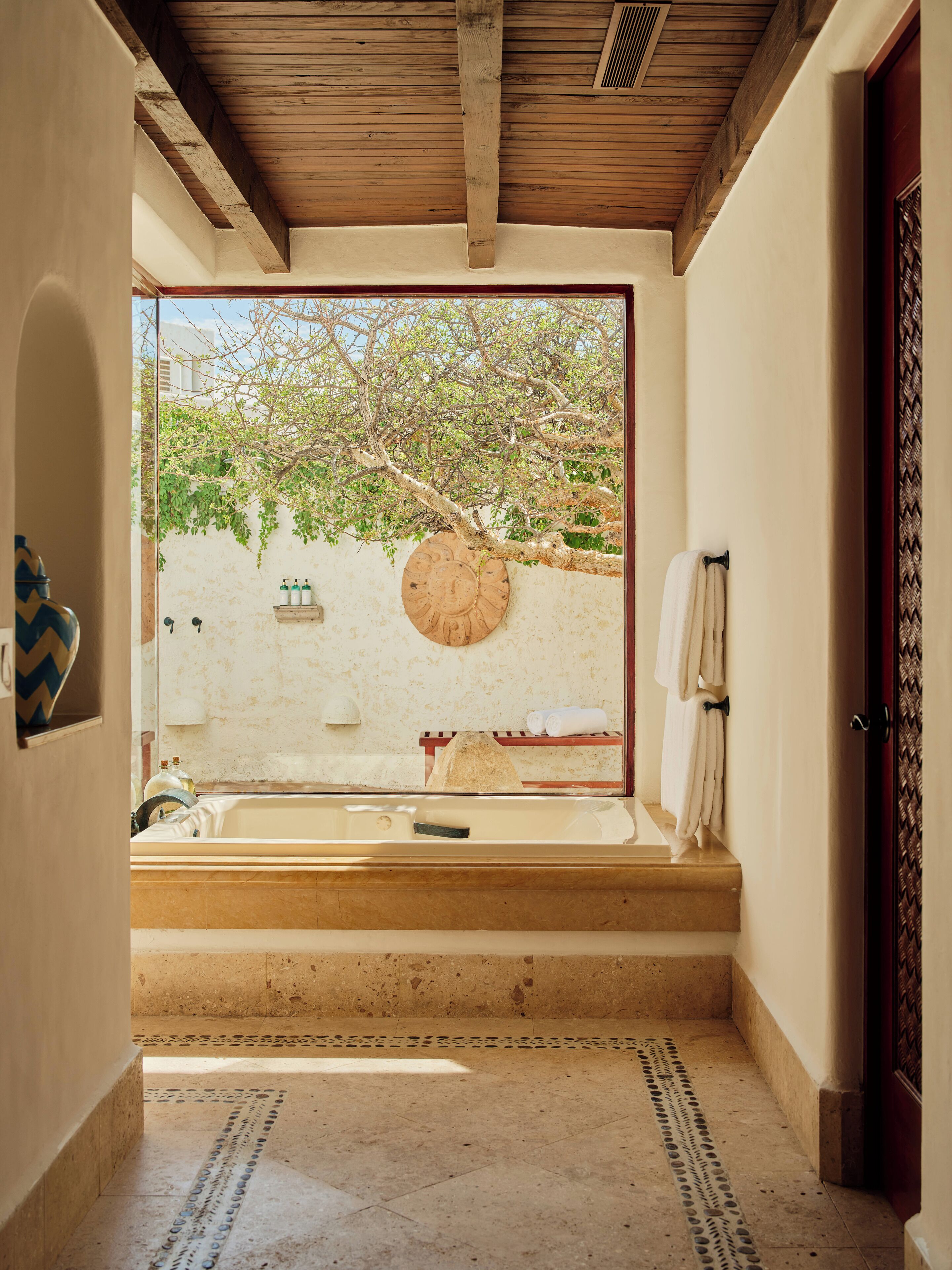 A bathroom with a bathtub, a towel rack, and a view of a tree and wall outside.