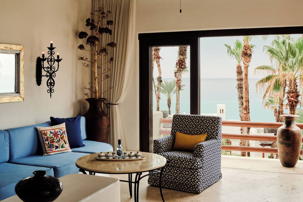 A room with a sofa, a patterned armchair, a small table, a vase, a candelabra, and a view of palm trees and the sea.