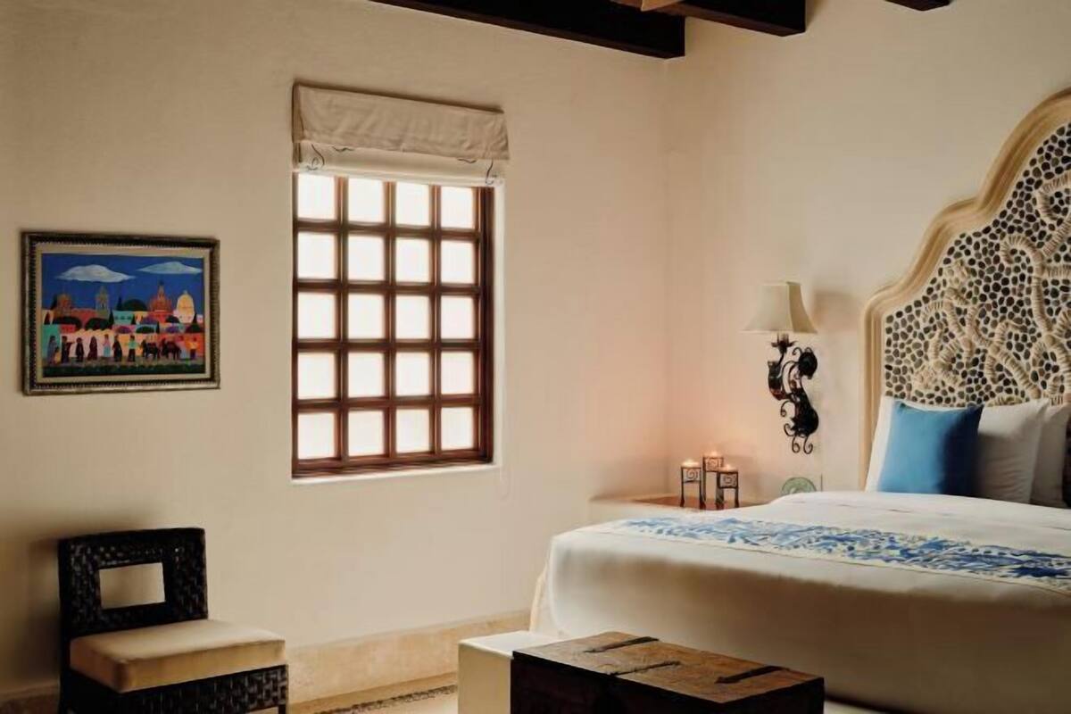A bedroom with a bed, a window with a view, a wall-mounted lamp, and a framed picture.