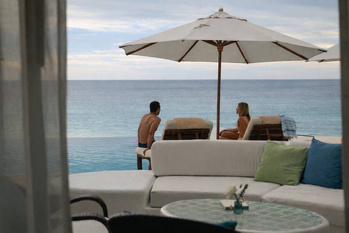 A view from a balcony overlooking the ocean, with a white sofa and an umbrella, and two people sitting on chairs.