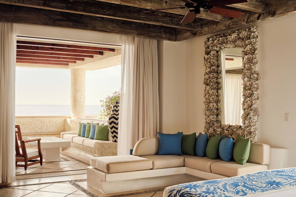 A room with a large bed, a sofa with pillows, a chair, a decorative mirror, and a view of the sea.