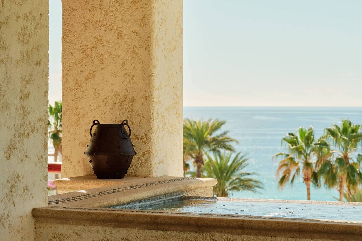 A stone fountain with a pot, overlooking a beach with palm trees and the ocean.