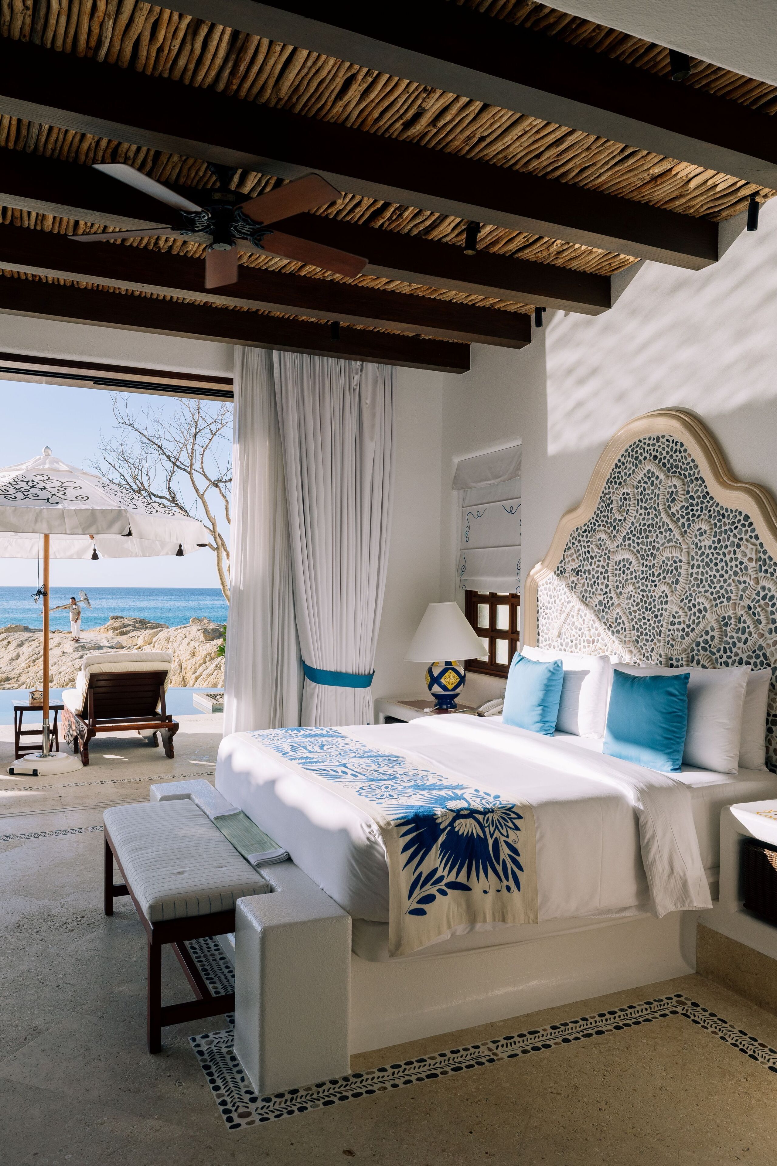 A bedroom with a bed, a bench, a nightstand, a lamp, and a view of the beach.