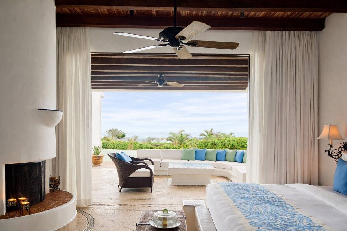 A hotel room with a curved sofa, a bed with a blue patterned bedspread, a ceiling fan, and a view of the outdoors.