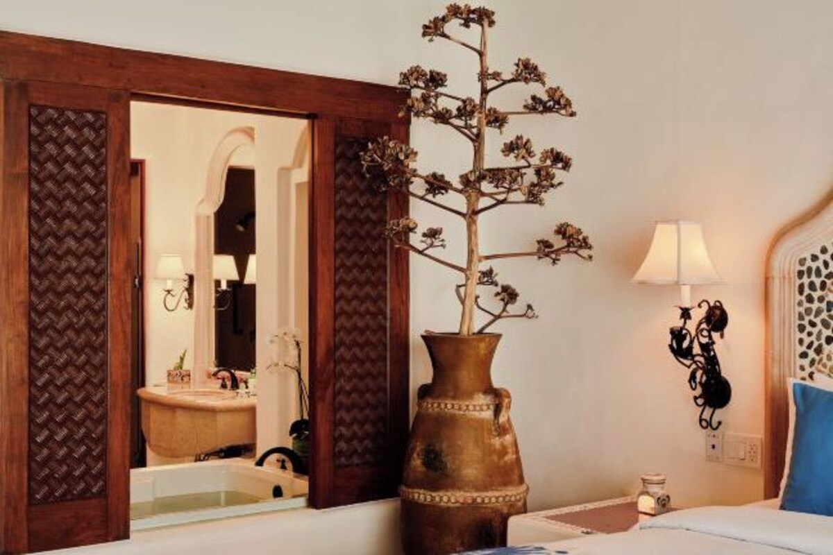 A hotel room with a large mirror, a wooden-framed door, a vase with dried flowers, a bed with a blue pillow, and a wall-mounted lamp.
