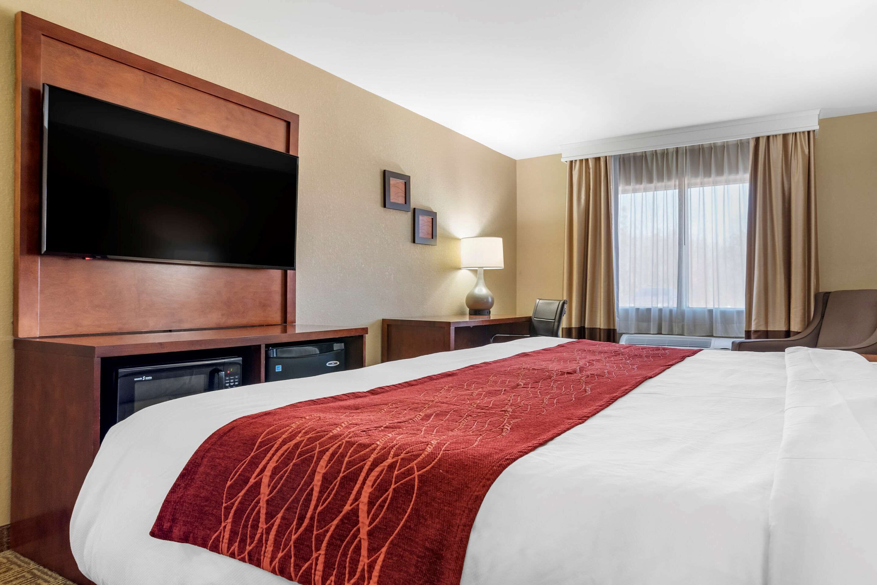 Habitación de hotel con una cama grande, un televisor de pantalla plana montado en la pared, un escritorio con lámpara y una ventana con cortinas.