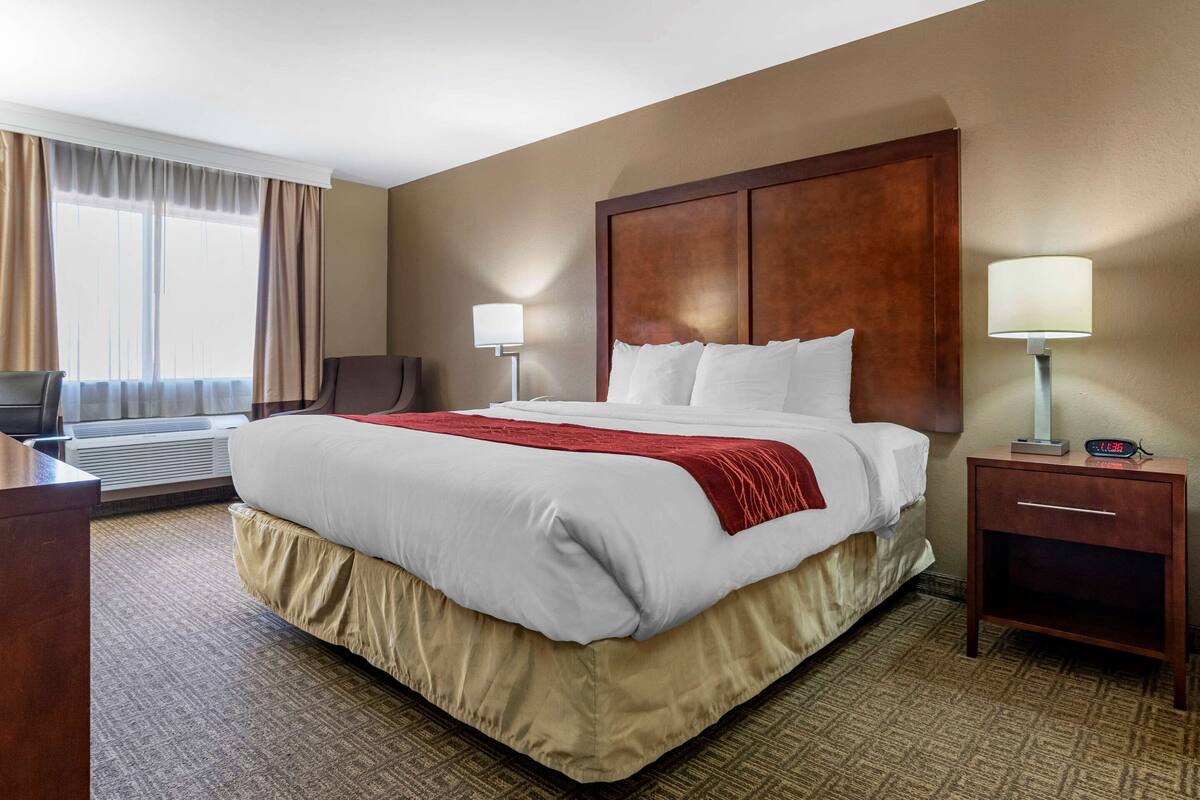 A hotel room with a large bed, two bedside lamps, a desk, and an air conditioning unit.