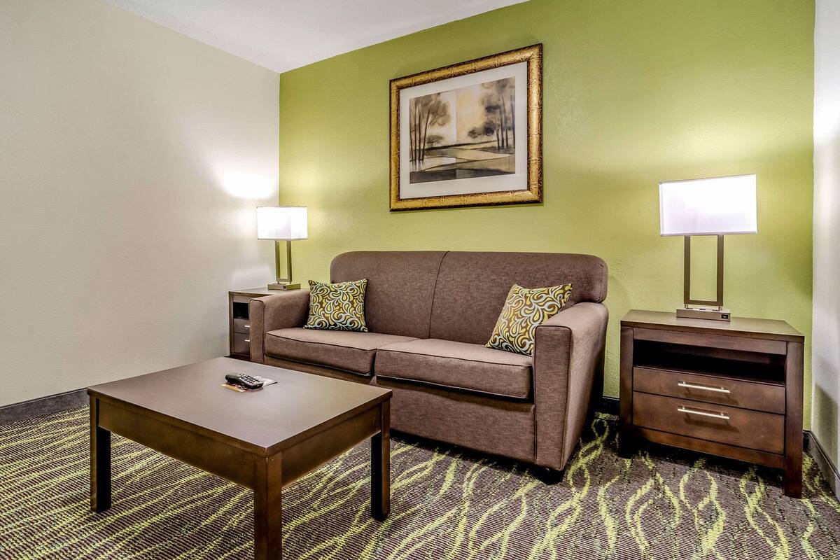 A hotel room with a brown sofa, a wooden coffee table, two bedside tables with lamps, and a patterned carpet.