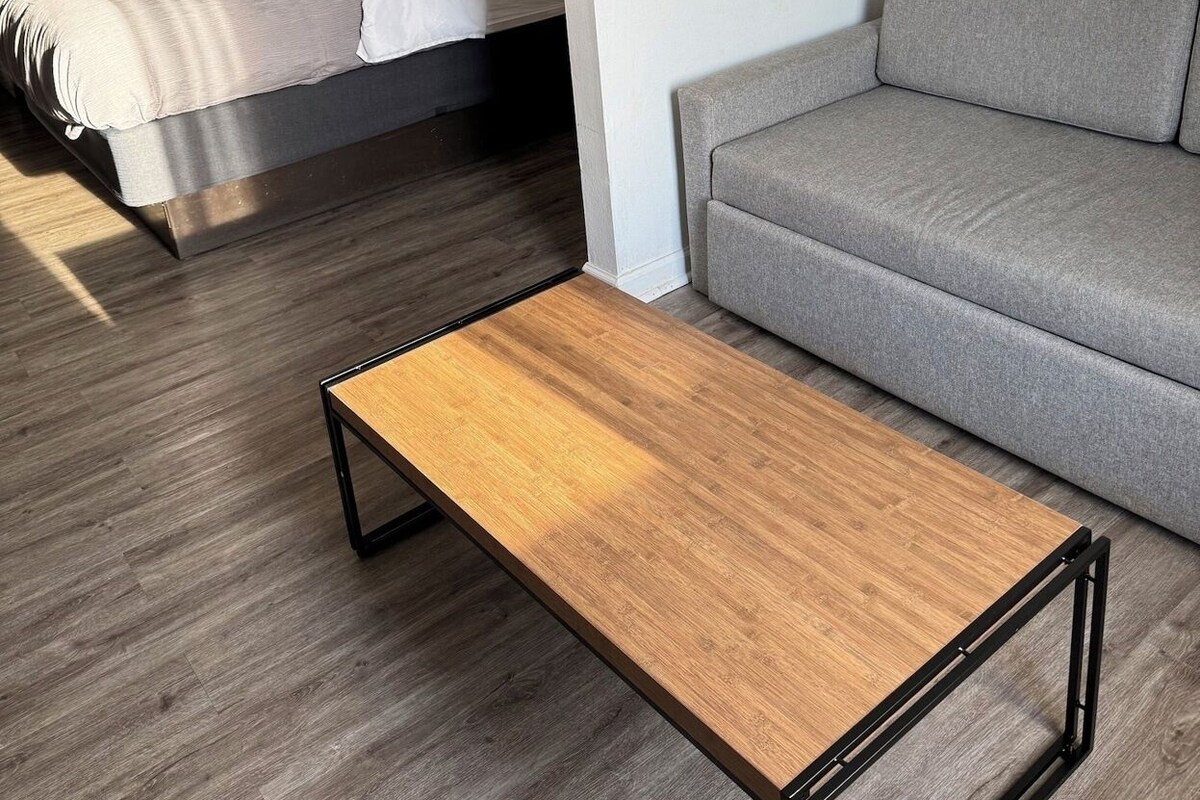 A modern hotel room with a bed, a grey sofa, a wooden coffee table, and a painting of a sunset over a pier.