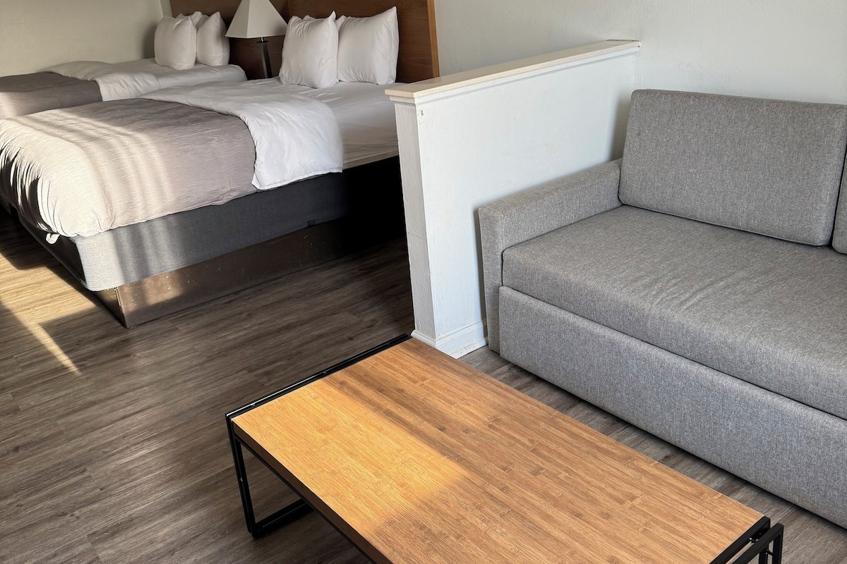 A modern hotel room with a bed, a grey sofa, a wooden coffee table, and a painting of a sunset over a pier.