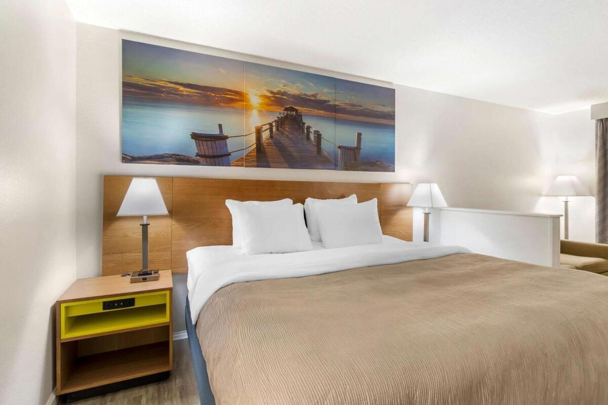 A hotel room with a bed, two bedside tables, a nightstand, a painting of a sunset over a pier, and a wooden headboard.