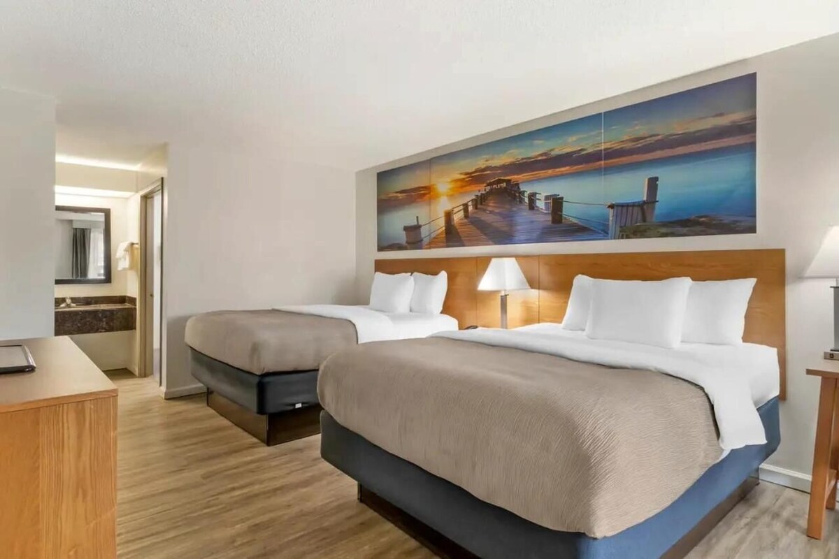 A hotel room with two beds, a desk, a mirror, and a large painting of a sunset over a pier.