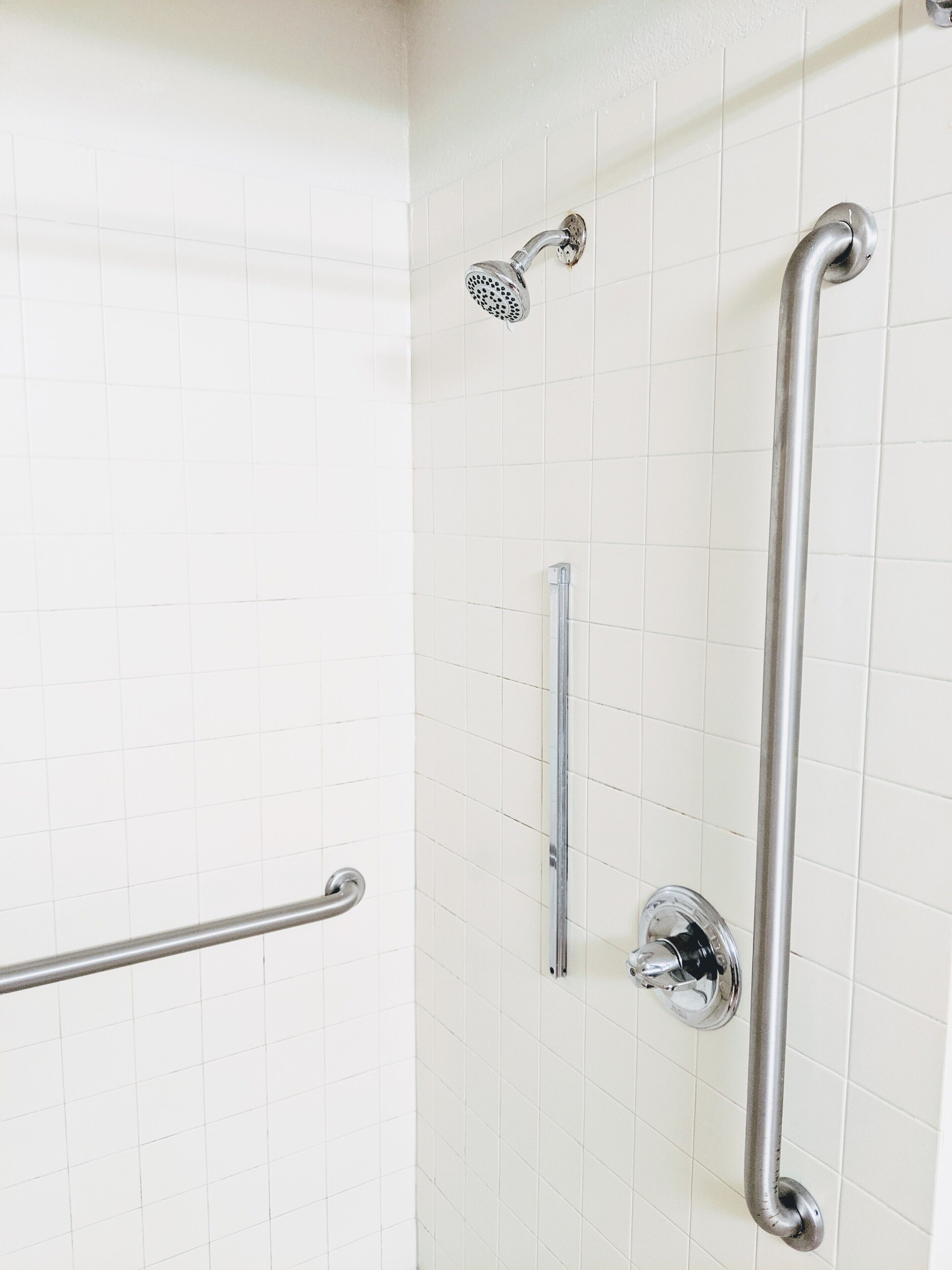 Une salle de bain avec des murs recouverts de carreaux blancs, une douchette à main, une barre d’appui et un robinet fixé au mur.