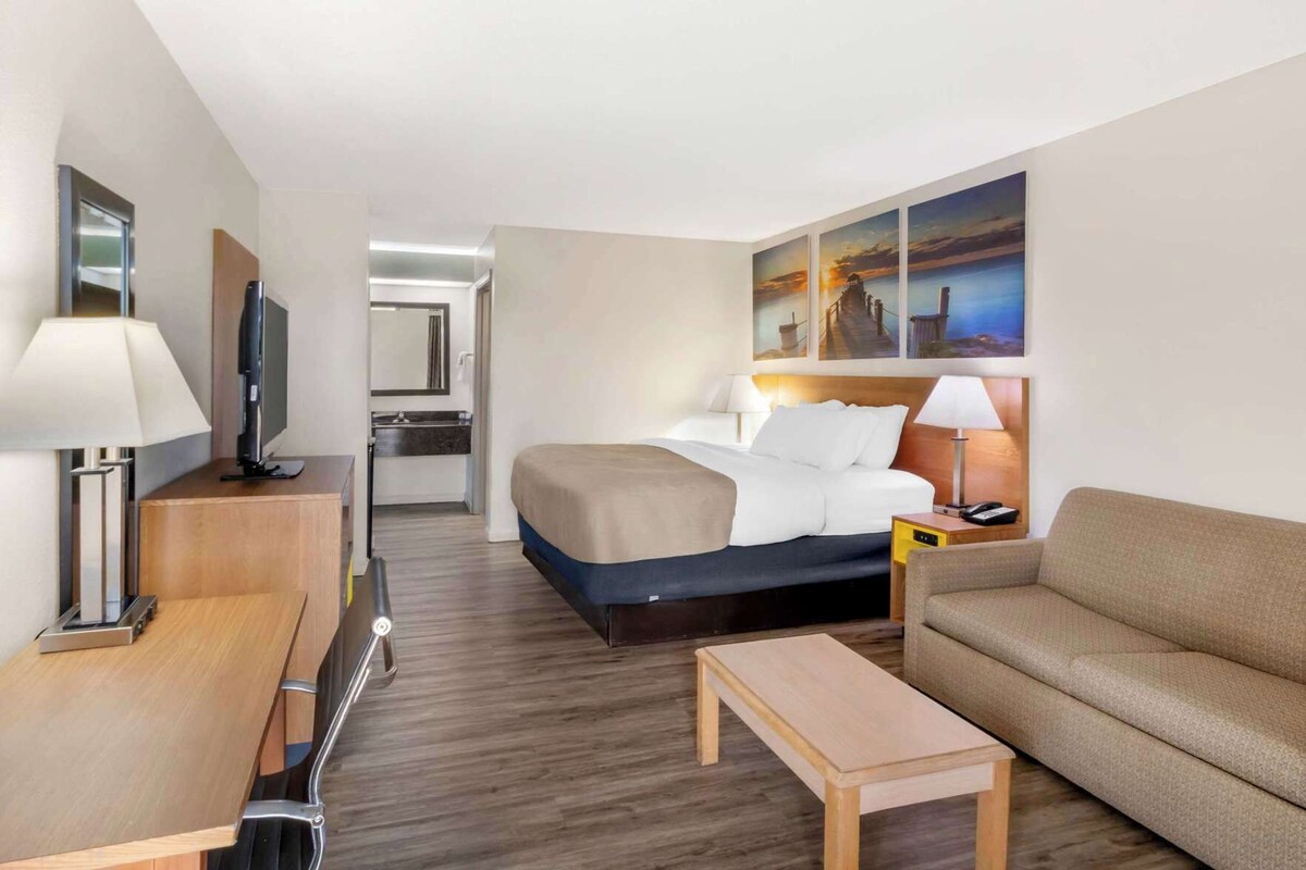 A hotel room with a bed, a sofa, a desk, a TV, and a painting on the wall.