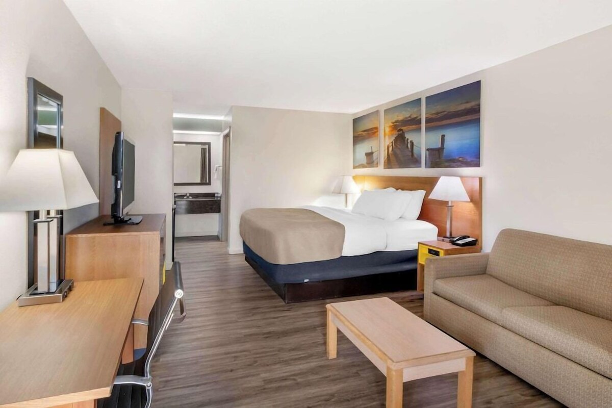 A hotel room with a bed, a sofa, a desk, a TV, and artwork on the wall.