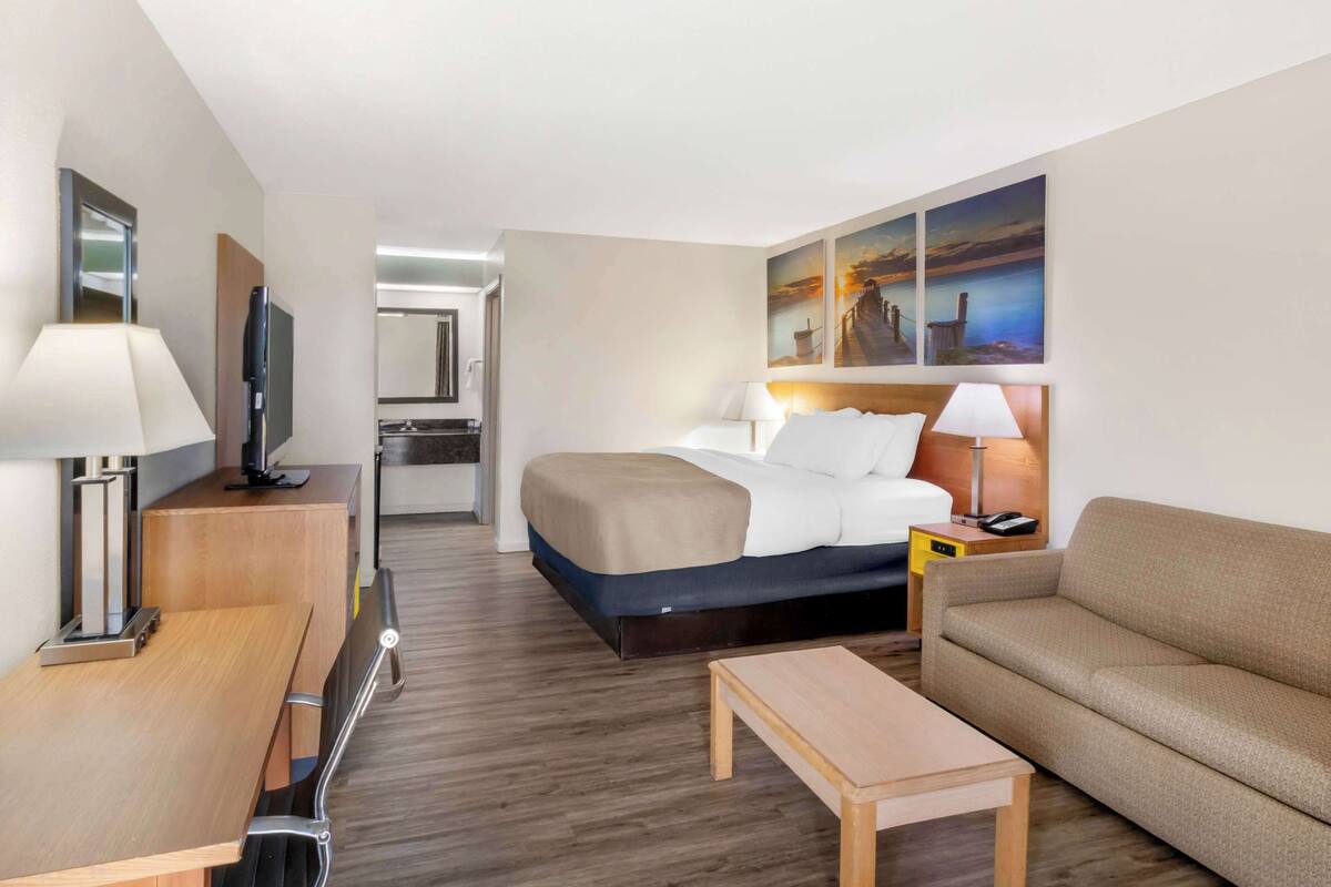 A hotel room with a bed, a sofa, a desk, a TV, and a painting on the wall.