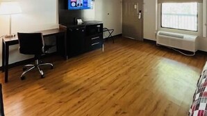 Ein kleiner, einfach eingerichteter Raum mit Holzboden, einem Schreibtisch mit Stuhl, einem an der Wand befestigten Flachbildfernseher und einem Fenster mit Blick nach draußen.
