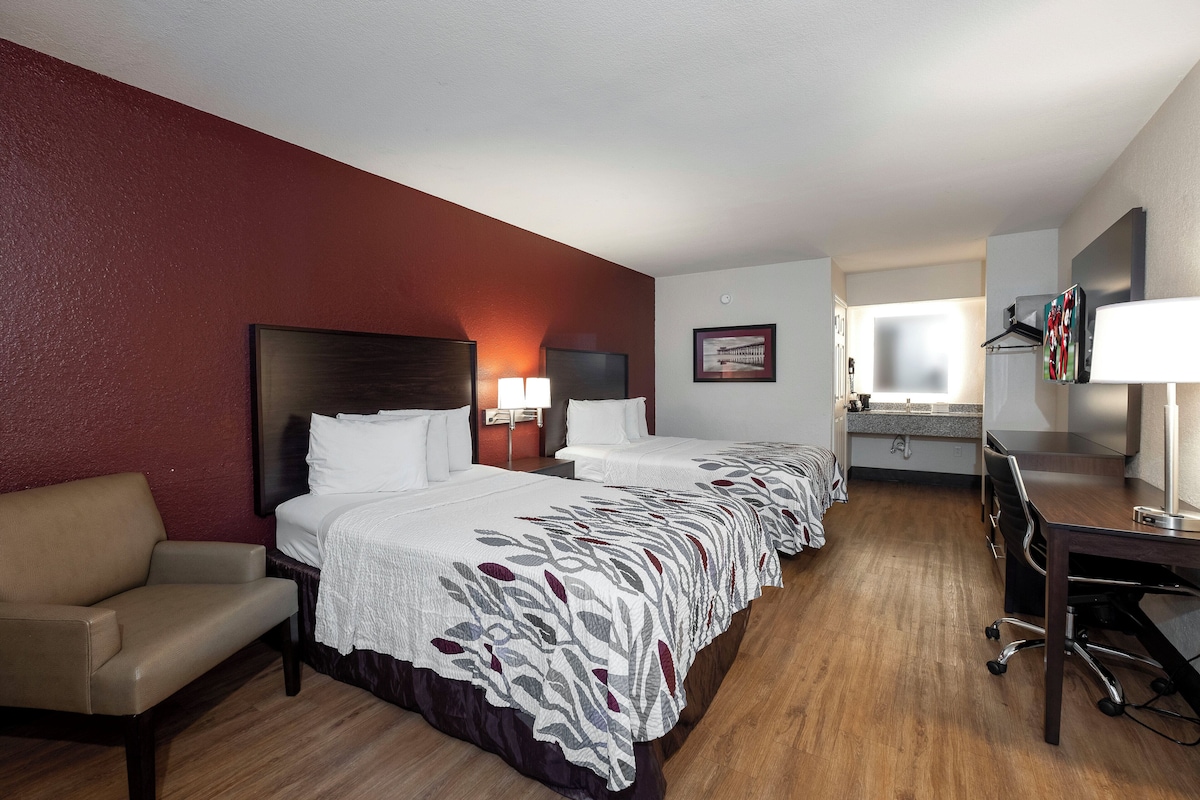 Een hotelkamer met twee bedden, een bureau, een stoel, een bureaulamp, een televisie en een ingelijste foto aan de muur.