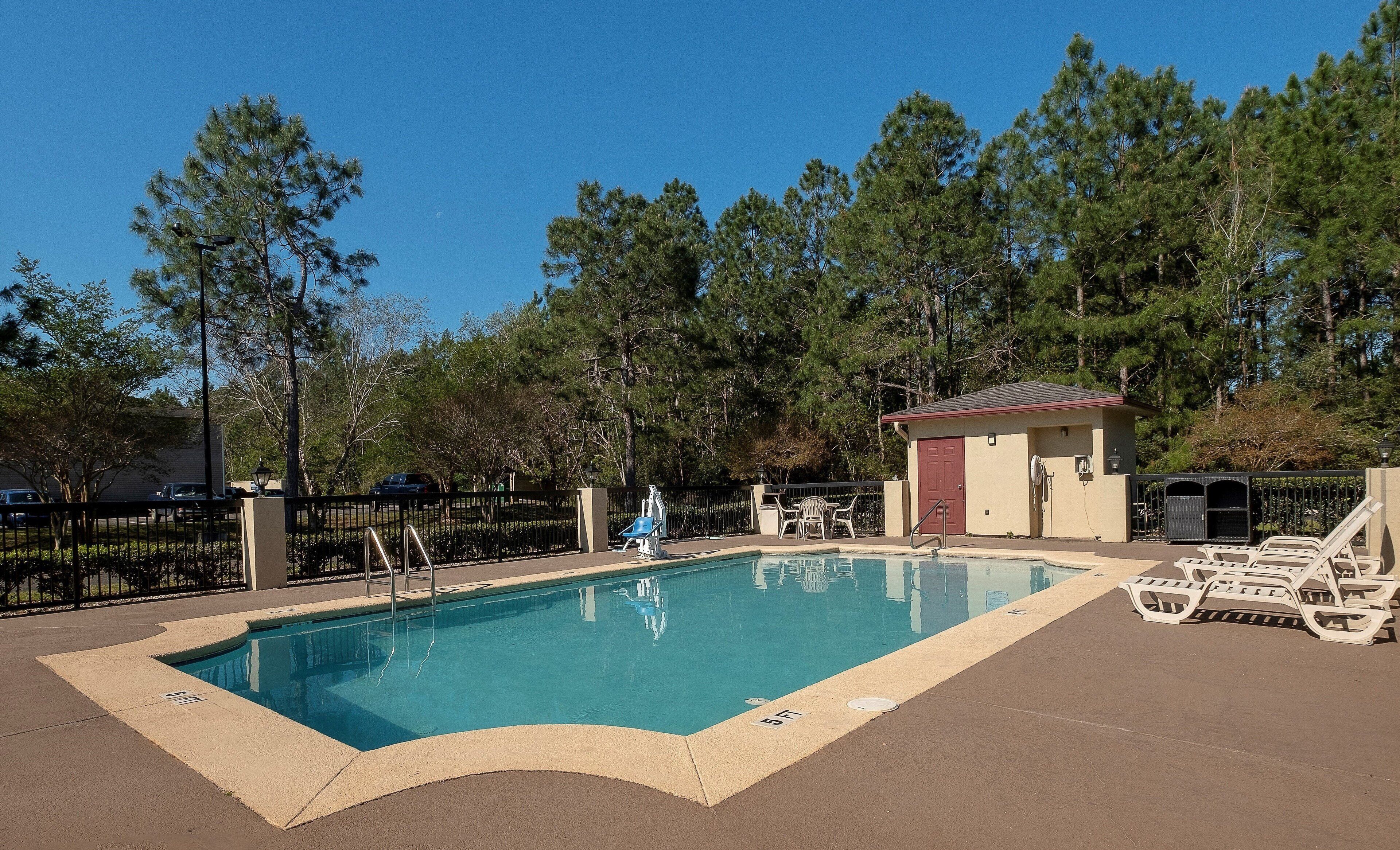 Piscina com cadeira de salva-vidas, espreguiçadeiras e um pequeno edifício, cercada por árvores.
