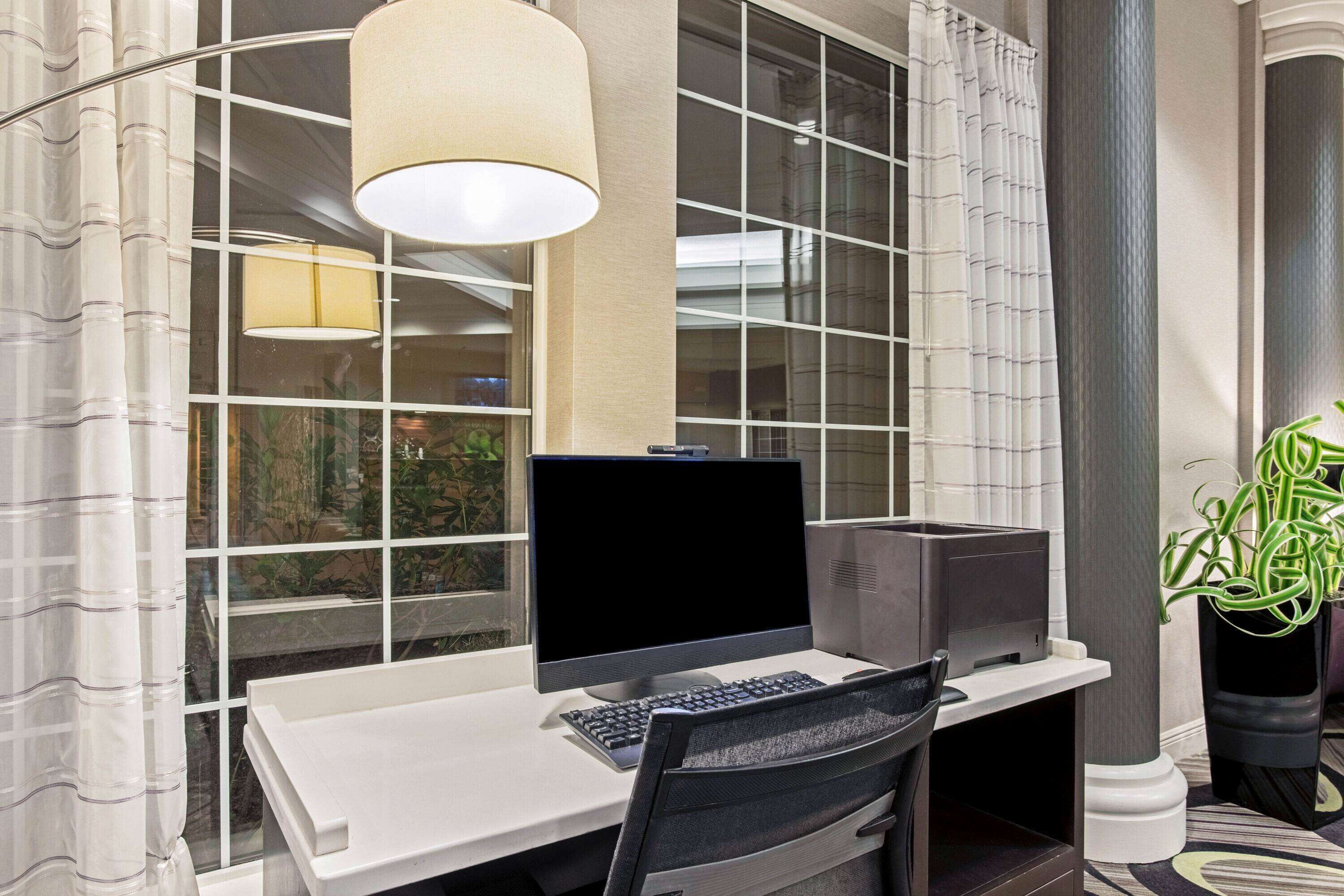 A modern office space with a desktop computer, a chair, a lamp, and a large window with curtains.