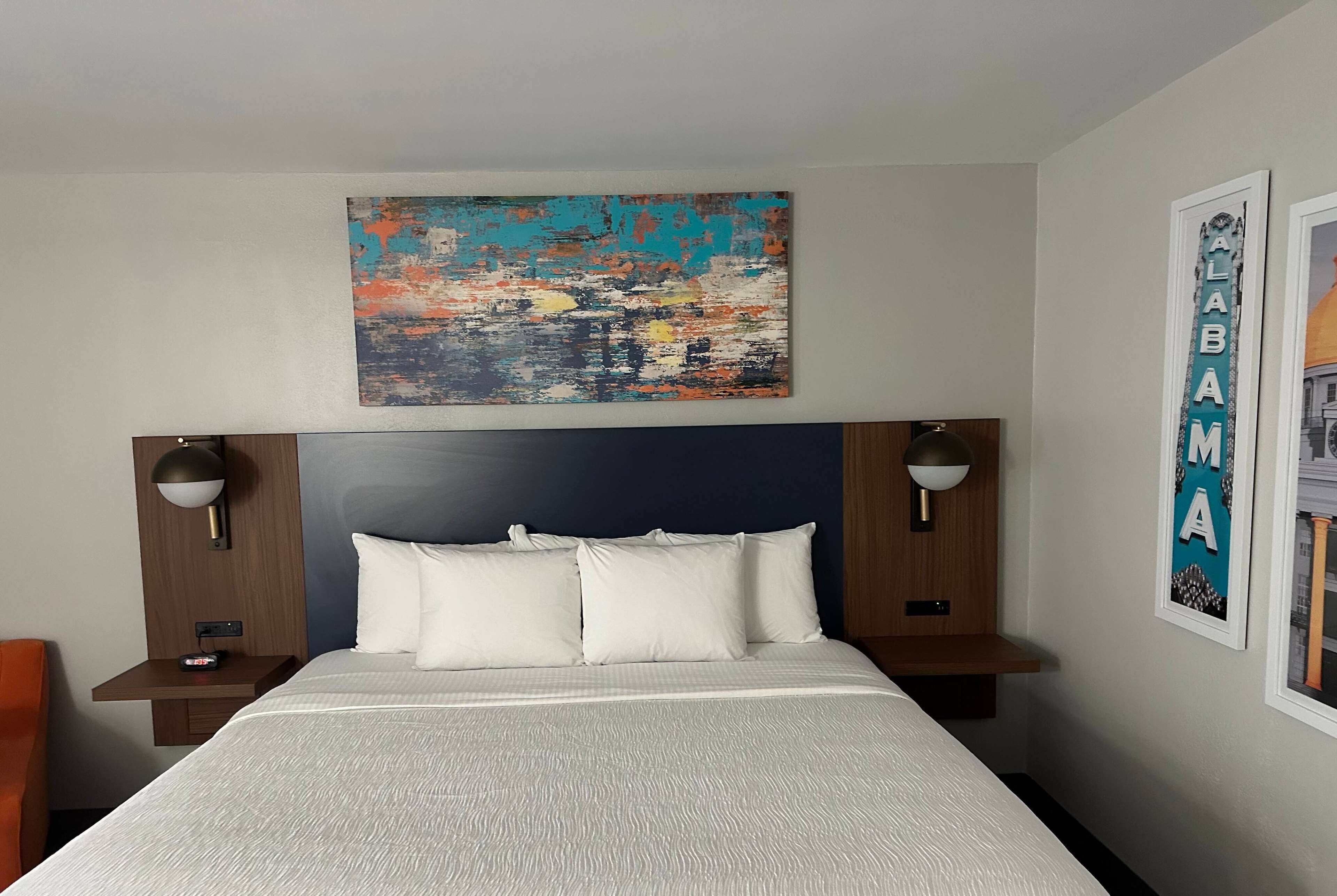 A hotel room with a bed, two bedside tables, a nightstand, a wall-mounted painting, and a framed sign.