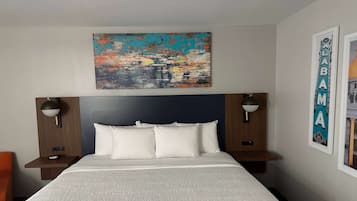 A hotel room with a bed, two bedside tables, a nightstand, a wall-mounted painting, and a framed sign.