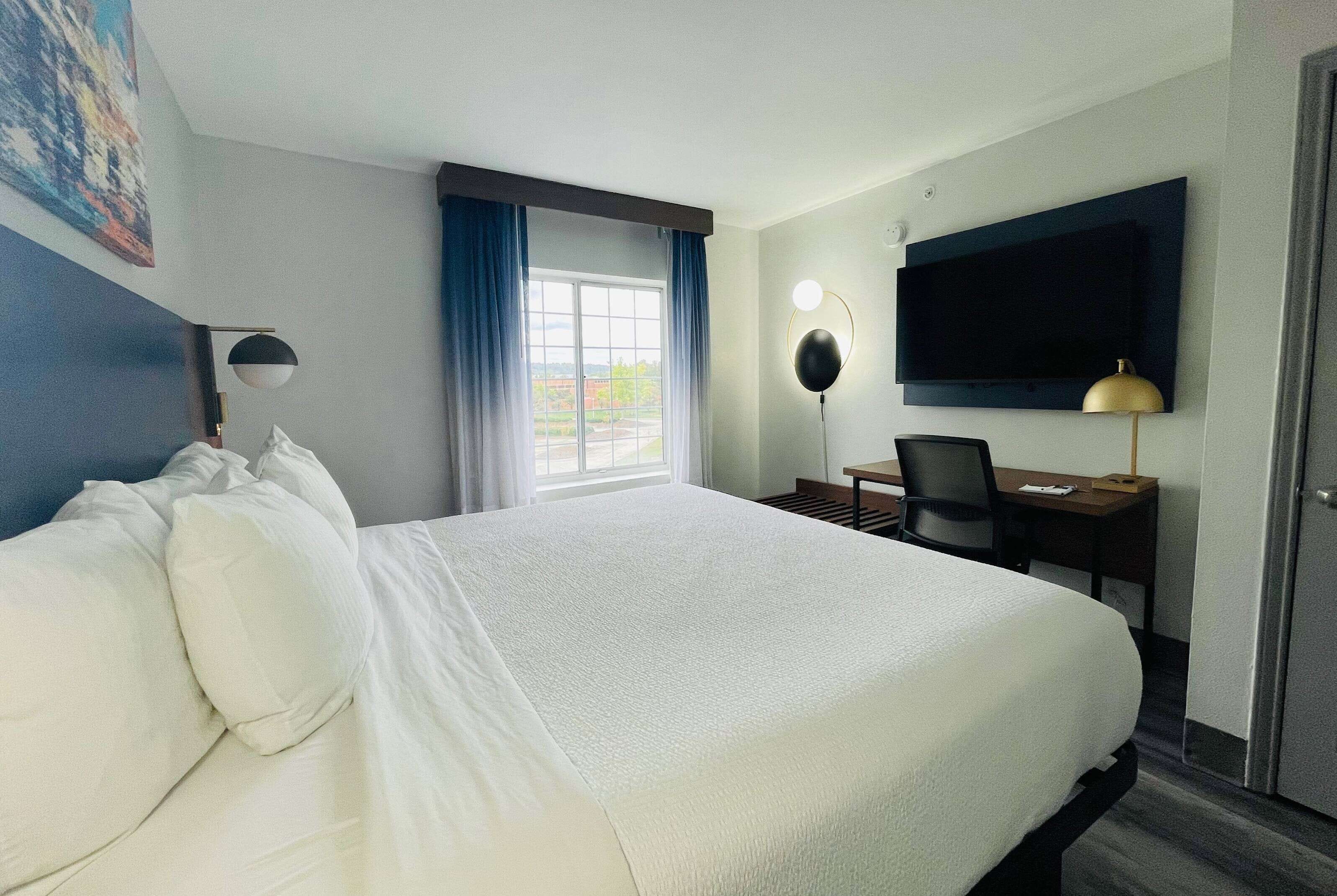 A hotel room with a large bed, a desk with a chair, a television, and a window with curtains.