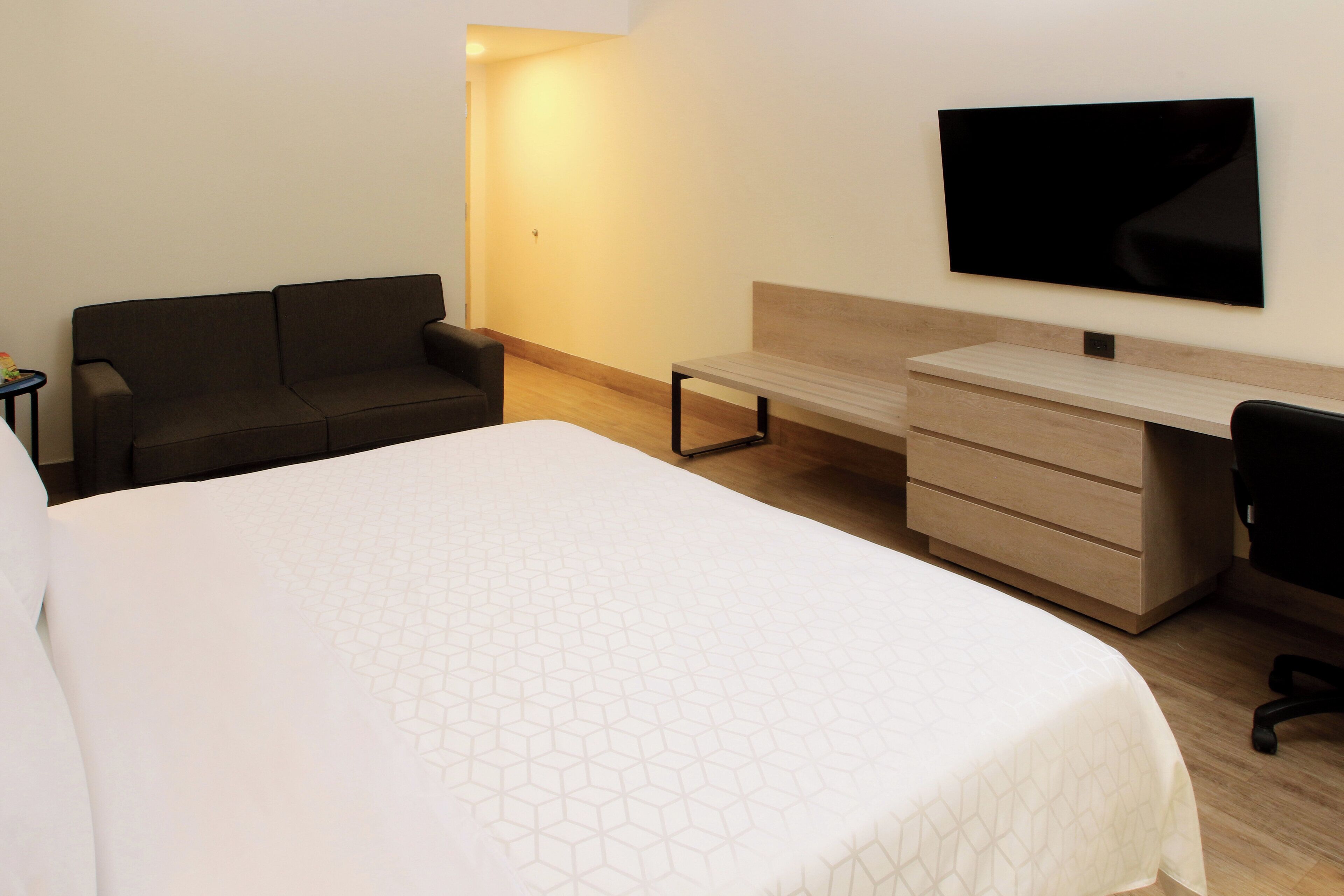 Quarto de hotel com uma cama grande, uma escrivaninha com gavetas, uma televisão de tela plana na parede e um sofá de cor escura.