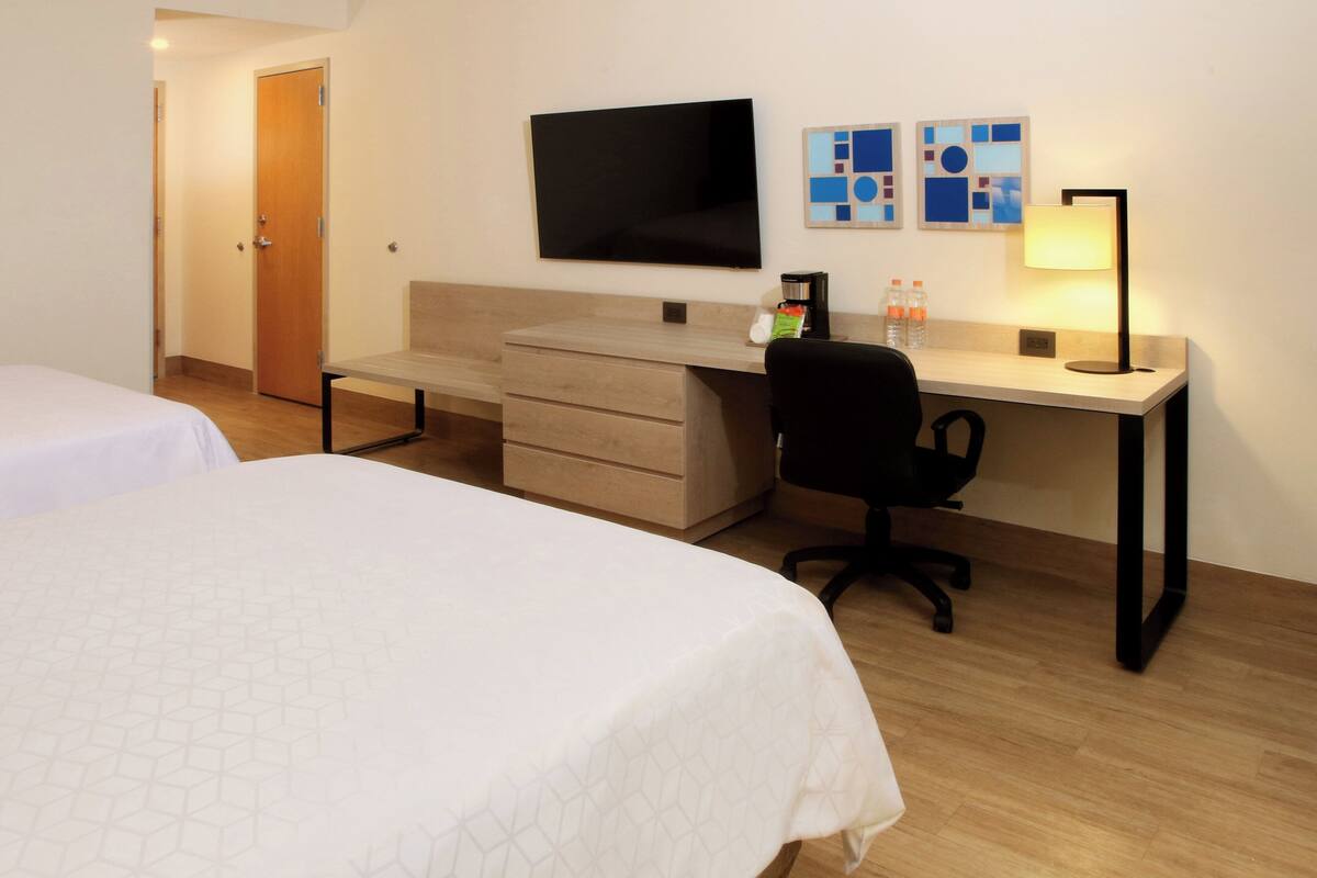 A hotel room with two beds, a desk with a chair, a flat-screen TV, and a lamp.