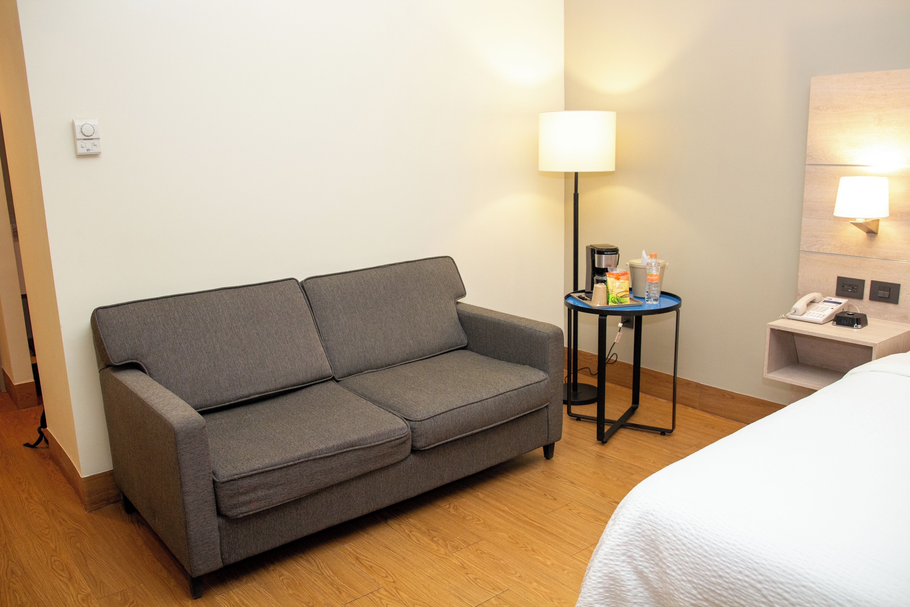 A hotel room with a grey sofa, a small side table with a lamp, a water bottle, and a phone.