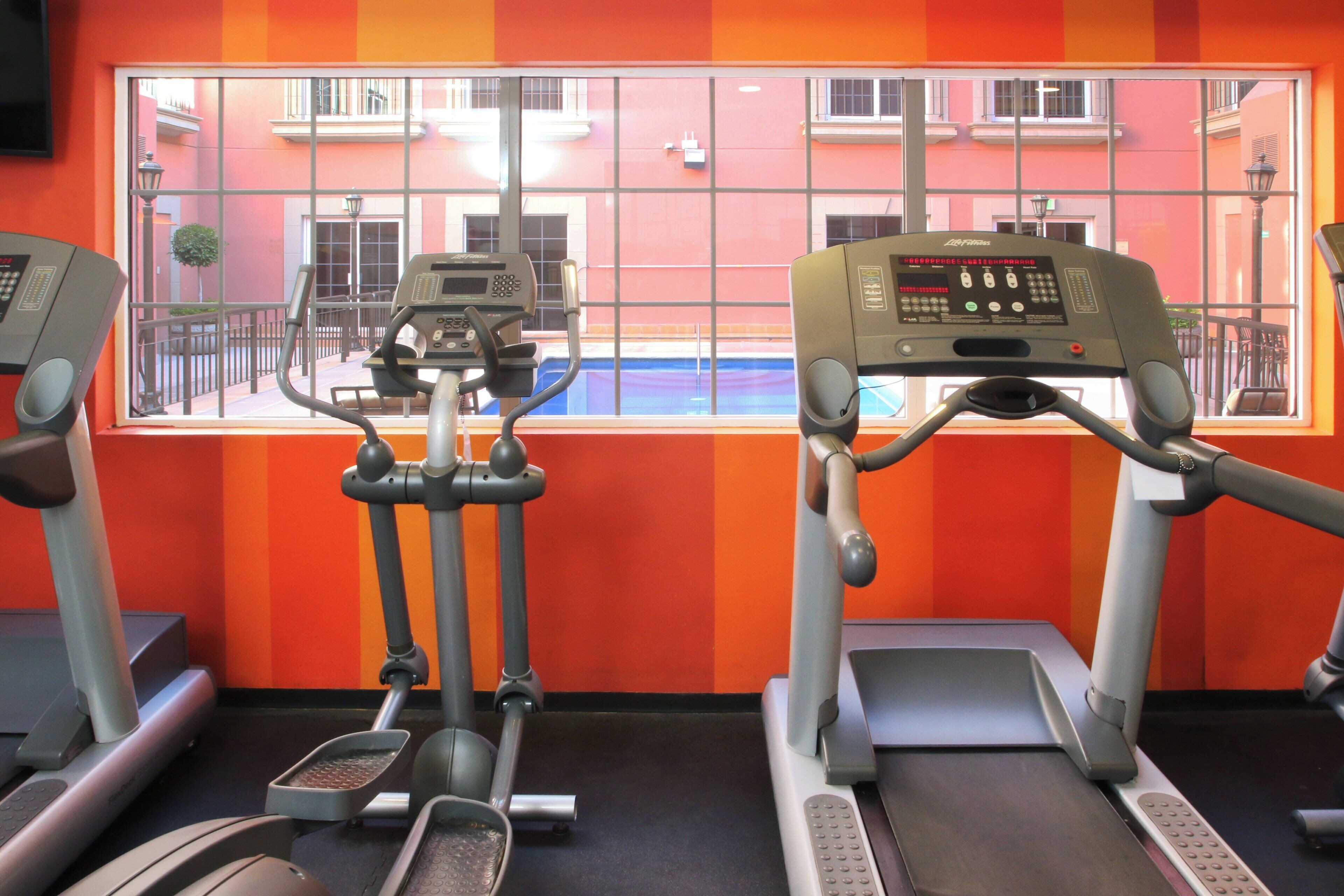 Un gimnasio con cintas de correr y elípticas, una ventana con vistas a edificios y una pared roja y naranja.