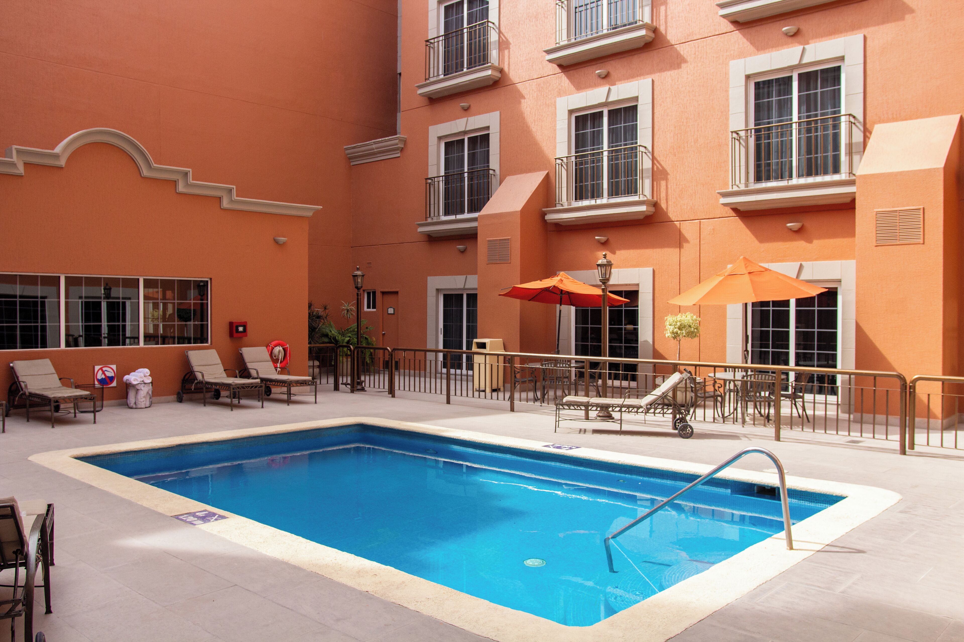 A swimming pool with lounge chairs and umbrellas, surrounded by a building with balconies and windows.