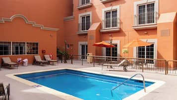 A swimming pool with lounge chairs and umbrellas, surrounded by a building with balconies and windows.