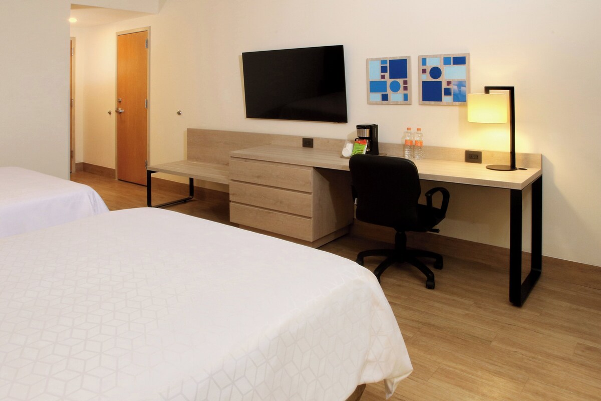 Habitación de hotel con dos camas, un escritorio con silla, un televisor de pantalla plana y una lámpara.