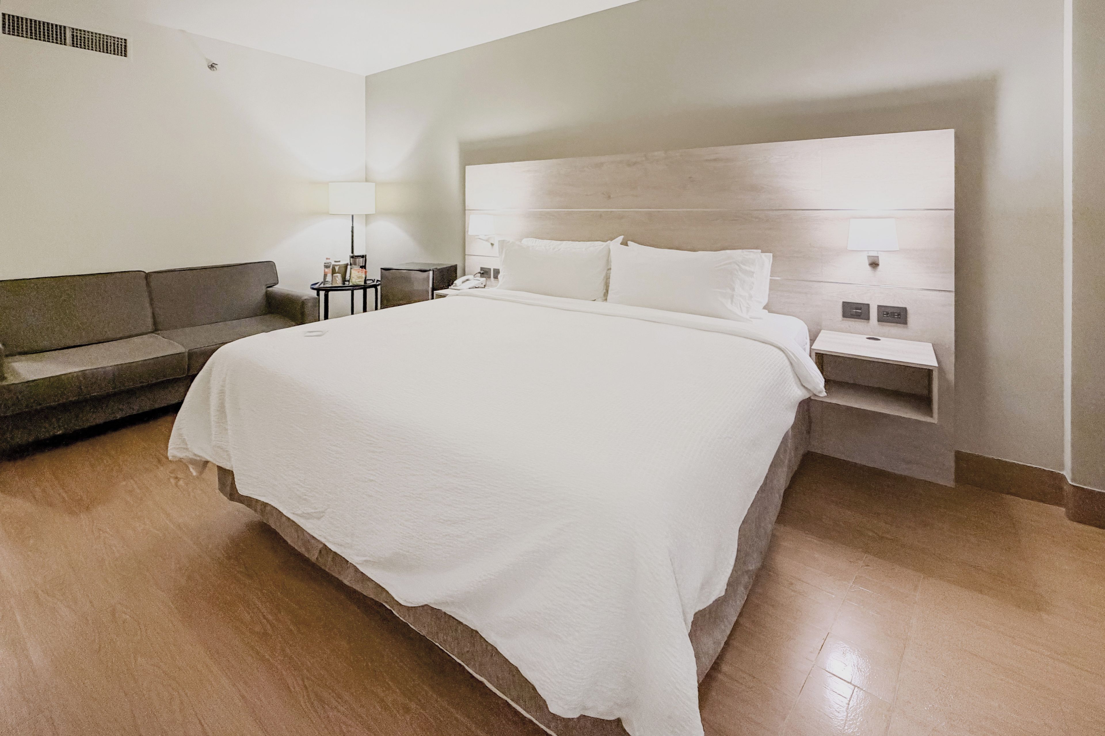 A hotel room with a large bed, a sofa, a bedside table with a lamp, and a nightstand with a phone.
