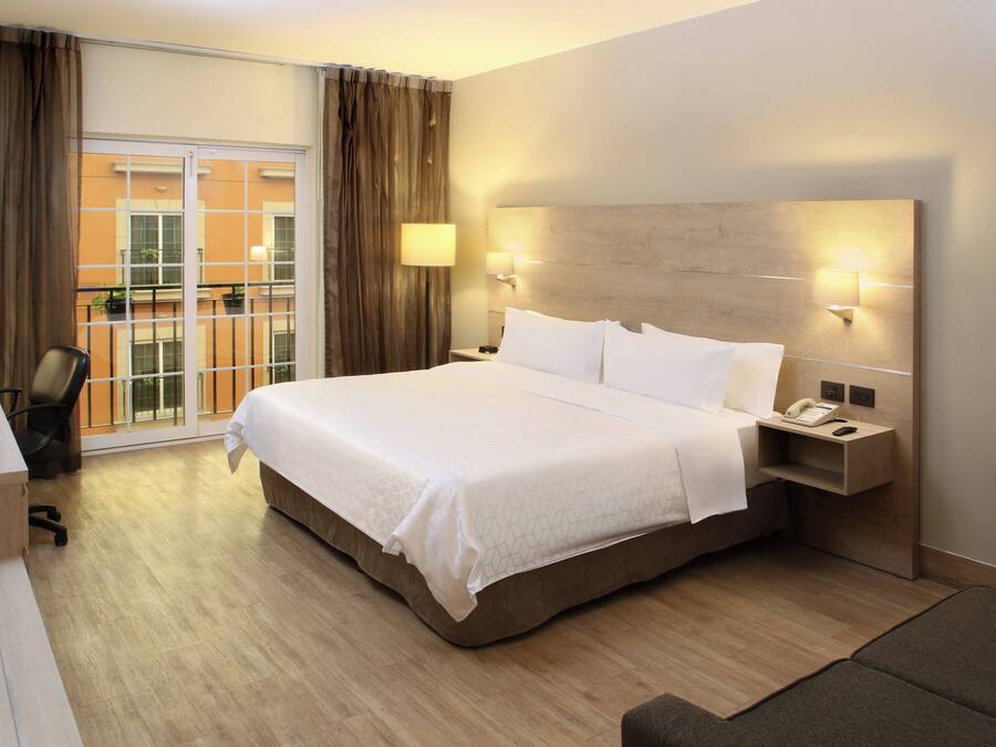 Habitación de hotel con una cama grande, un escritorio, una silla y vistas al balcón exterior.