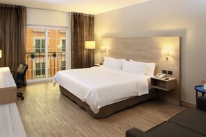 Habitación de hotel con una cama grande, un escritorio, una silla y vistas al balcón exterior.