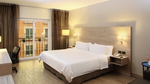 Habitación de hotel con una cama grande, un escritorio, una silla, una mesita de noche con teléfono y vistas al balcón exterior.