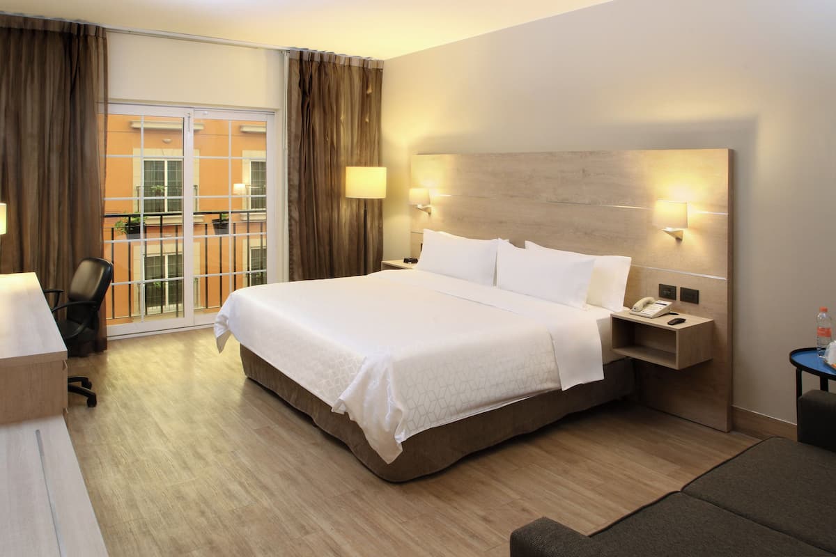 A hotel room with a large bed, a desk, a chair, a nightstand with a phone, and a view of the outside balcony.