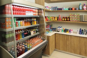 Un refrigerador de bebidas repleto de varias opciones, una estantería con aperitivos y una encimera con artículos de aseo personal.