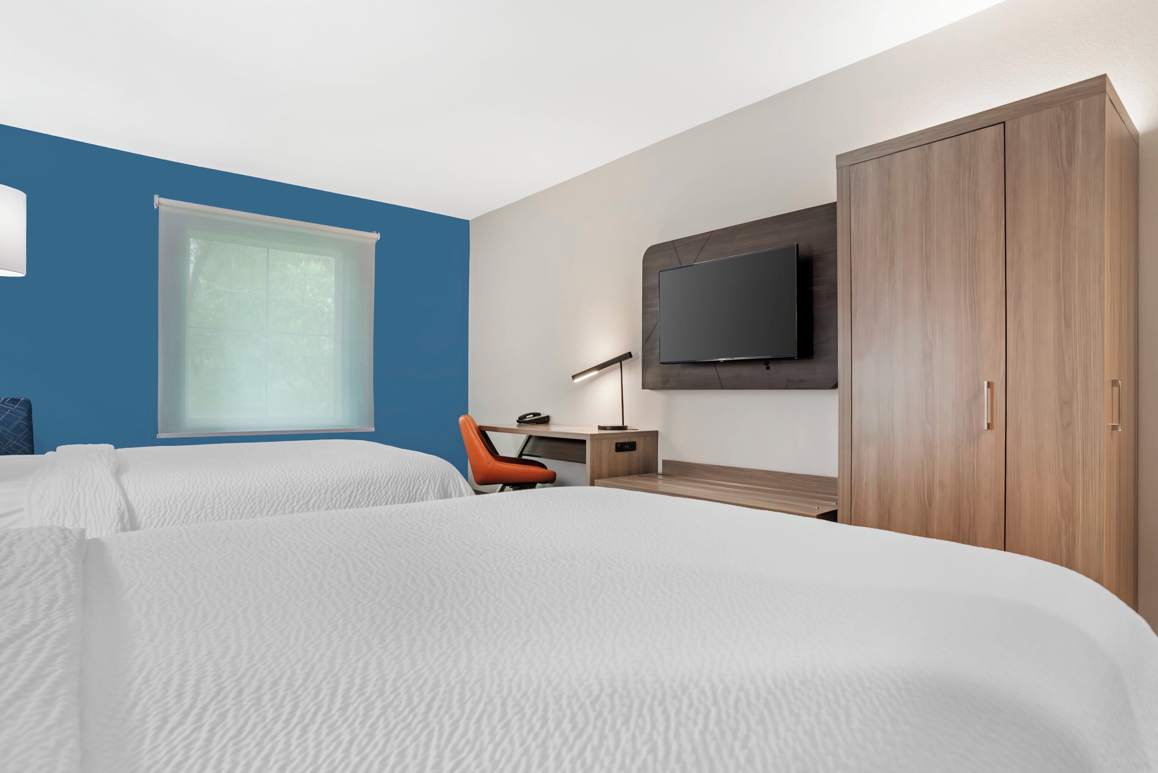 Habitación de hotel con dos camas, un escritorio con silla, un televisor montado en la pared y un armario.